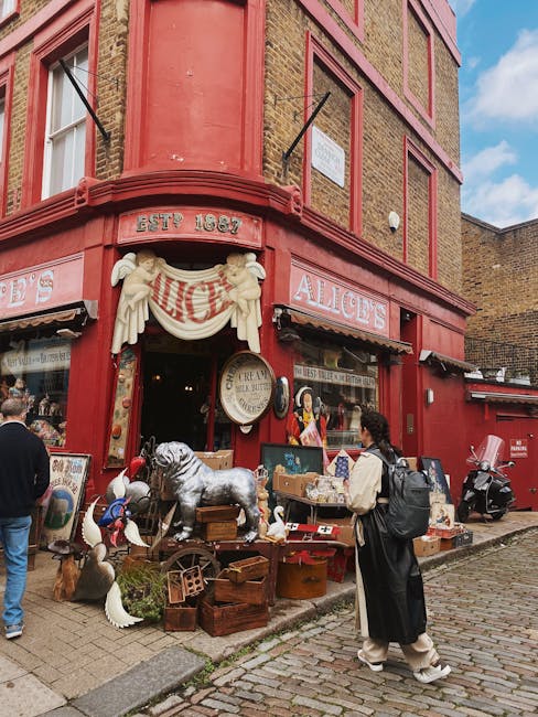 This image depicts the exterior of Alice's antique shop situated on a corner building with a red brick facade and decorative brickwork in Islington. The shop's entrance features a hanging sign and a vintage-style awning. Outside, various retro and vintage items are displayed on tables and wooden crates, including a silver dog statue, white ceramic ducks, and framed artwork. The setting is lively with pedestrians walking along the cobblestone street, one woman in a black coat and backpack observing the display. The surrounding area appears clean and well-maintained, with natural daylight illuminating the scene. Despite the outdoor setting, this image emphasizes the importance of general cleanliness and maintenance of such vintage retail spaces, aligning with the themes of surface cleaning and sanitation that [COMPANY_NAME] offers for commercial properties like this shop.