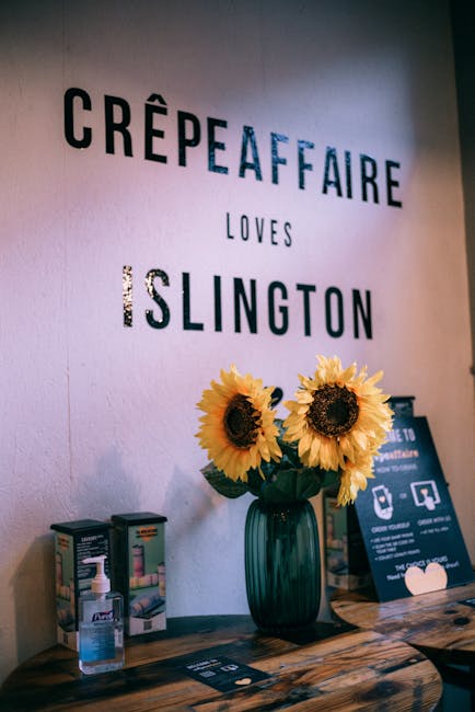 Interior view of a tea or crepe shop wall featuring a decorative sign that reads 'Crêpe Affaire Loves Islington' in bold, black letters with a blue accent. Below the sign, there's a dark green glass vase holding a bouquet of vibrant yellow sunflowers placed on a wooden table or counter. To the left of the vase, a small hand sanitizer dispenser and some product boxes are visible, indicating cleanliness and hygiene measures. The surface of the wooden table appears to be well-maintained, clean, and polished, reflecting good surface cleaning practices. The lighting is soft, highlighting the freshness of the flowers and the cleanliness of the display area, fitting within a setting that benefits from professional cleaning by Islington Cleaners, as referenced on the page about Angel cleaners guide Chapel Market Islington.