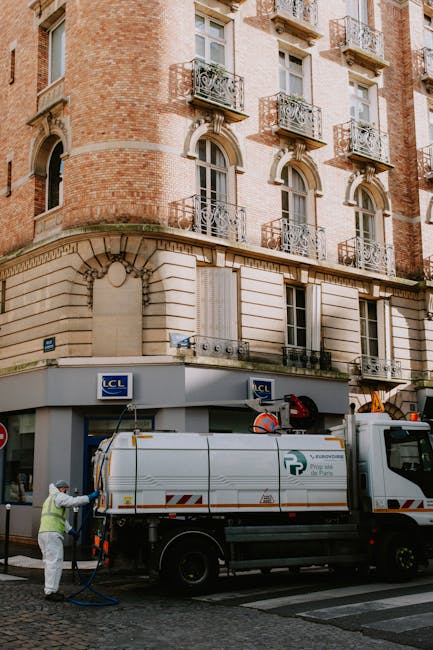 A worker dressed in protective clothing is operating a cleaning machine connected to a large vacuum truck on a cobblestone street in front of a historic multi-story building with a cream-colored stone lower façade and red brick upper floors. The building features decorative wrought iron balconies, arched window frames, and large windows. The scene highlights urban surface cleaning and sanitisation processes, demonstrating professional domestic or commercial cleaning services as offered by Islington Cleaners. The lighting is natural, with daylight illuminating the scene, emphasizing the cleanliness and maintenance activities around the building's exterior.