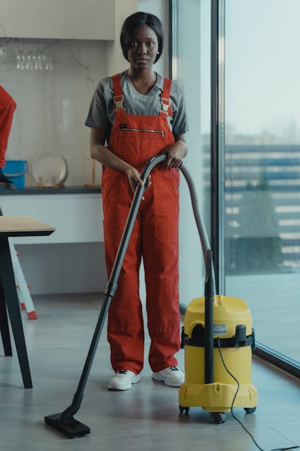 A professional cleaner, a woman of African descent, is in a modern kitchen with a large glass sliding door leading to a balcony. She is dressed in a grey t-shirt and red overalls, holding the handle of a yellow commercial vacuum cleaner with an extended metal hose and a flat cleaning nozzle. The kitchen features a white countertop with a blue dish rack and glassware on the shelves above, along with a wooden table partially visible to the side. The floor is made of large, light grey ceramic tiles that are visibly clean and polished, reflecting natural light coming through the glass door, which reveals a blurred outdoor balcony. The scene highlights surface cleaning, deep cleaning, and sanitisation processes typical of domestic cleaning in an Islington property, performed by Islington Cleaners to maintain hygiene and overall cleanliness.